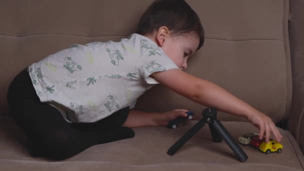Small brunette boy is playing with plastic cars on the couch moving them and arranging — Stock Video