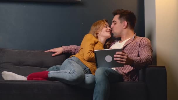 Joven pareja encantadora tumbada en el sofá y sonreír besándose usando una tableta — Vídeo de stock