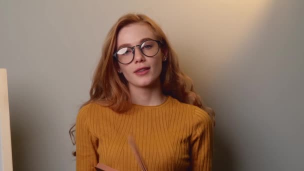 Freckled ginger woman is thinking a bout something while speaking on a gray wall holding a book and smile — Stock Video