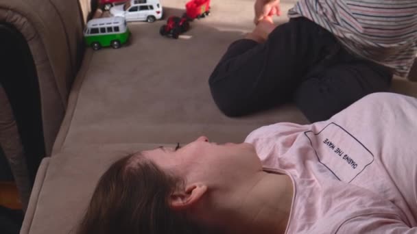 Mãe deitada no sofá e sorrindo enquanto seu filho está brincando com ela e abraçando — Vídeo de Stock