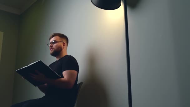 Blanke bebaarde man heeft online video klassen leraar anderen iets uit een boek terwijl zitten op een grijze studio muur met licht — Stockvideo