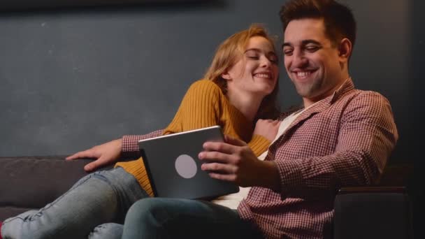 Caucásico hombre está utilizando una tableta, mientras que su mujer pelirroja con pecas lo besa en la mejilla y sonrisa acostado en un sofá — Vídeo de stock