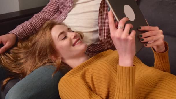 Ruda pani z piegami uśmiecha się leżąc na kolanach kochanków i używając tabletu — Wideo stockowe