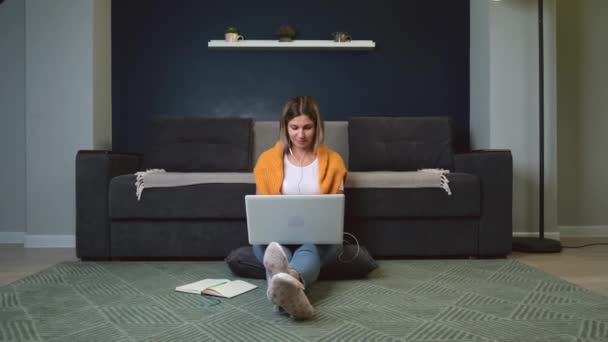 Caucasian woman is working at the laptop on the floor while using earphones and keeping a book opened — Stock Video