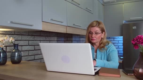 Elfoglalt idős nő iszik egy kávét, miközben távolról dolgozik a laptopnál a konyhában otthon. — Stock videók