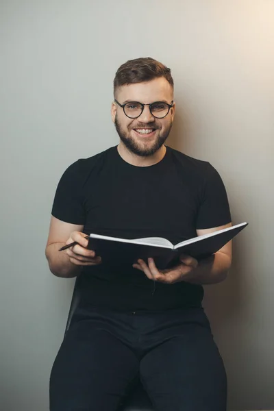 Homem barbudo sorrindo para a câmera em uma parede de estúdio cinza segurando um livro e vestindo óculos — Fotografia de Stock