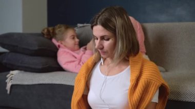 Beyaz bir anne yerde çalışırken kızı arka planda bir kanepede uzanmış telefonda oynuyor.