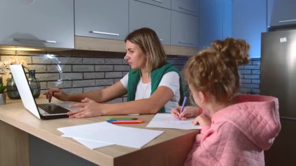 Biała matka pracuje nad laptopem w kuchni, podczas gdy jej córka coś do niej rysuje. — Wideo stockowe