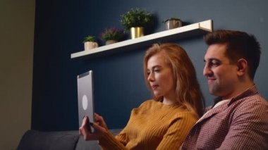 Kafkasyalı çift kanepede bir tablet kullanıyor. Gülümsüyor ve sevgililer gününü birlikte geçiriyorlar.
