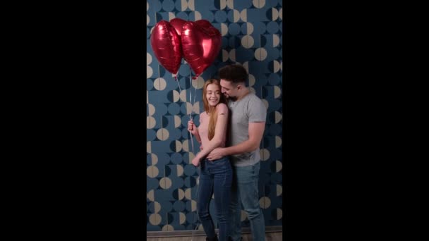 Кавказская пара обнимается на голубой стене, держа воздушные шары и улыбаясь — стоковое видео