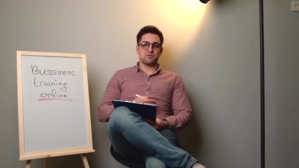 Homem caucasiano com óculos está explicando aulas de negócios na câmera sentada em uma cadeira perto da placa — Vídeo de Stock