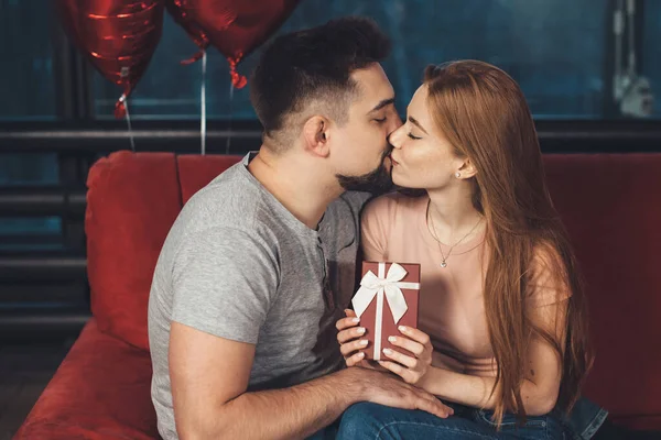 백인 생강 여인이 빨간 소파에 앉아 있는 발렌타인 데이에 선물을 받고 연인에게 키스를 하고 있습니다. — 스톡 사진