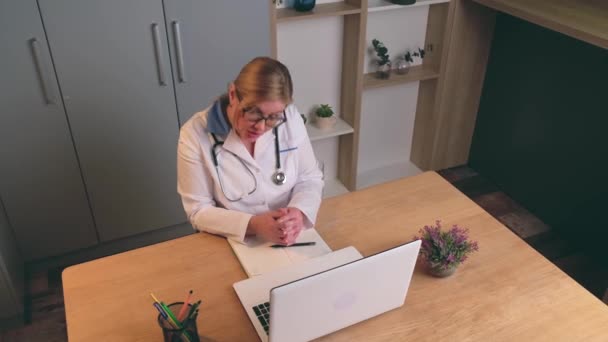 Vídeo vista superior de um médico loiro caucasiano falando on-line no laptop com o paciente no escritório — Vídeo de Stock