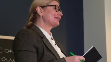 Okuldaki kıdemli sarışın öğretmen elinde bir kitapla tahtanın yanındaki dersi açıklarken gülümsüyor.