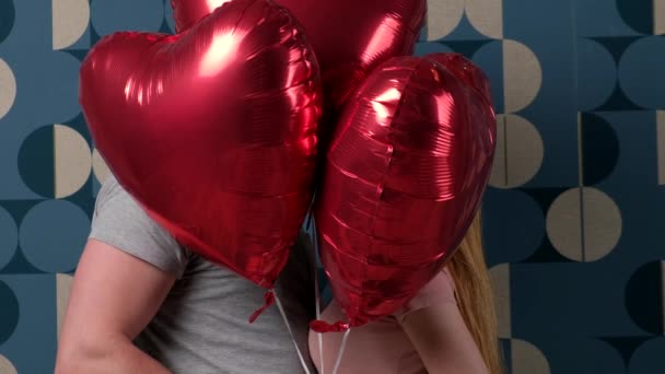 Caucásico pareja besándose en un azul pared mientras se esconde detrás de rojo globos — Vídeo de stock