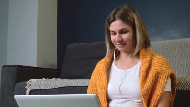 Mulher caucasiana está navegando no chão com um laptop enquanto ouve música em fones de ouvido — Vídeo de Stock