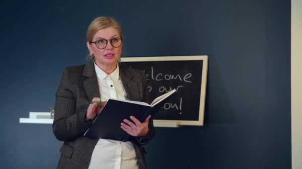 Старший учитель кавказского языка объясняет урок перед классом возле доски с книгой и в очках с формальной одеждой — стоковое видео
