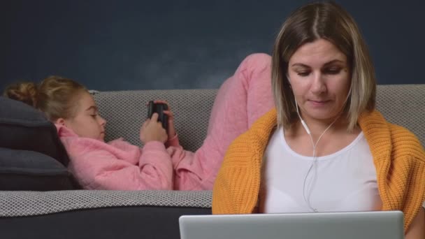 Blondynka pracuje zdalnie przy laptopie, podczas gdy jej córka leży na kanapie i używa telefonu. — Wideo stockowe
