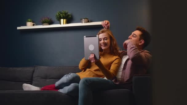 Młoda para leżąca na kanapie, relaksująca się i obejmująca oglądając coś na tablecie cyfrowym — Wideo stockowe