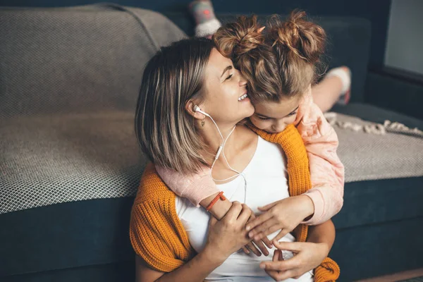 Caucásica hija abrazando a su mamá mientras ella está escuchando música en los auriculares y sonrisa — Foto de Stock