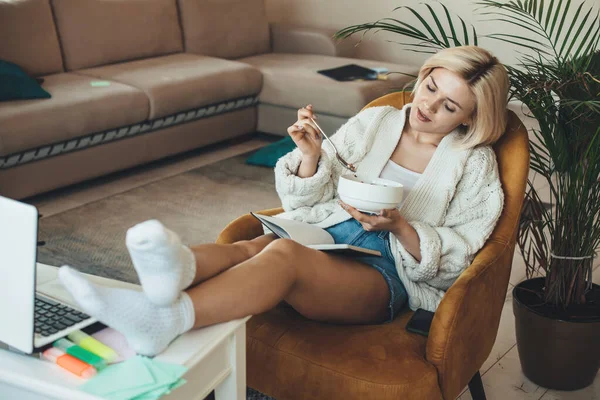 Krásná blondýnka sedí v křesle a dívá se na něco na notebooku, zatímco jí cereálie — Stock fotografie