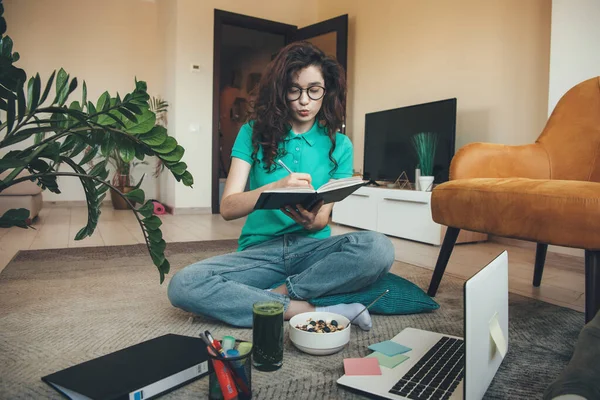 Кавказская девушка с кудрявыми волосами проводит онлайн урок на полу за ноутбуком во время еды зерновых и свежего зеленого сока — стоковое фото