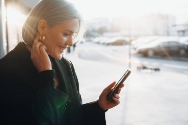 Mavili beyaz bir kadın telefonda konuşurken kulağına taktığı takılara dokunuyor.