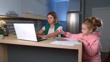 Beyaz bir kadın mutfakta dizüstü bilgisayar üzerinde çalışırken kızı onun yanına yaklaşıyor.