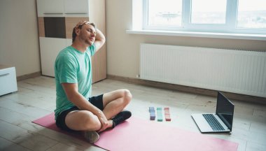 Esneyen beyaz adam spor kıyafetleriyle yerde oturuyor ve spor salonunda dizüstü bilgisayar kullanıyor.