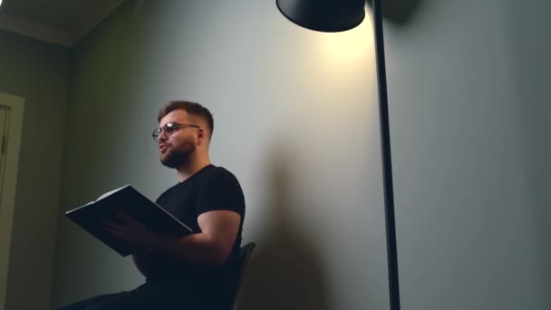 Белый мужчина с бородой проводит онлайн-курсы на серой стене студии, разговаривая с камерой из книги — стоковое видео