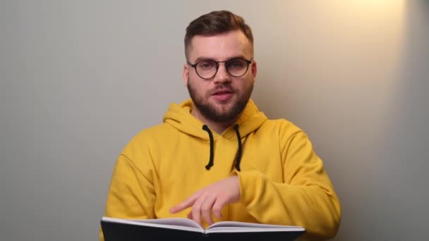 Caucásico hombre con gafas y barba está sosteniendo un libro y hablando en una videollamada — Vídeos de Stock