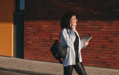 Beyaz bir öğrenci okuldan sonra yürüyor ve bir dizüstü bilgisayarla telefonda konuşuyor.
