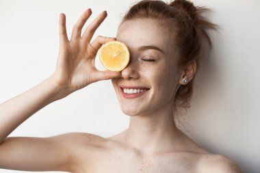 Kızıl saçlı sevimli bir kadın beyaz bir stüdyo duvarındaki limonla gözlerini kapatıyor.