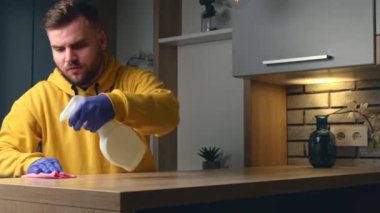 Sarı kazaklı sakallı beyaz adam antibakteriyel spreyle masayı temizliyor.