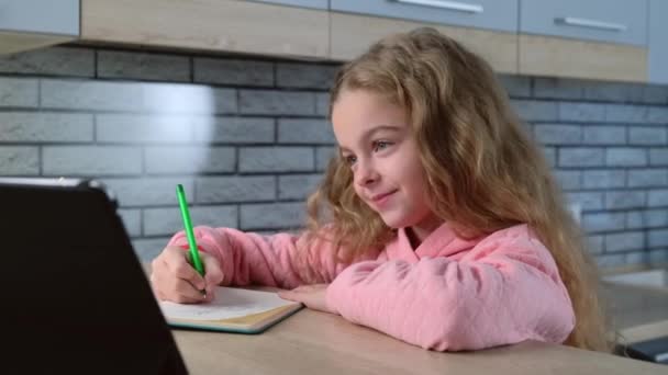 Kaukasisch klein meisje lacht terwijl het hebben van online lessen op een tablet en het opschrijven van het onderwerp — Stockvideo