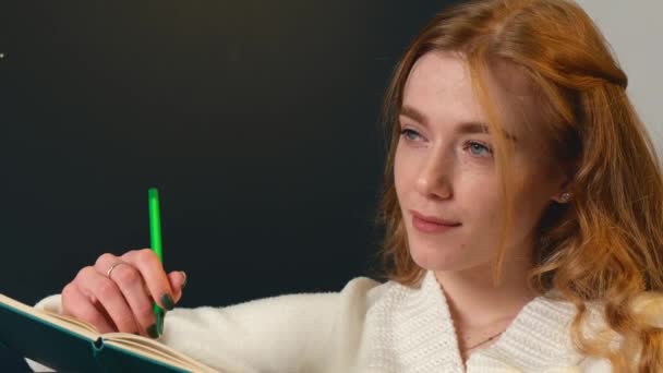 Mujer caucásica pensativa con pecas y el pelo rojo está escribiendo algo en un copybook — Vídeos de Stock