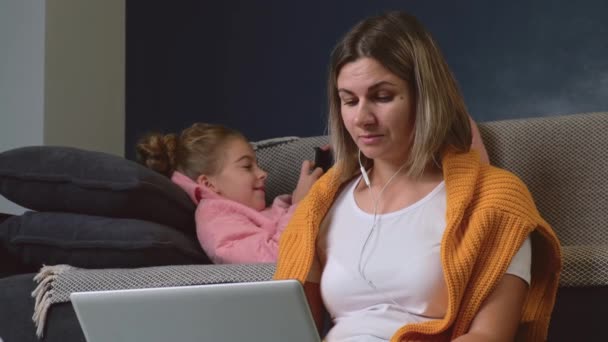 Rušné běloška matka pomocí notebooku a sluchátek, zatímco její dcera hraje na mobilu za ní — Stock video