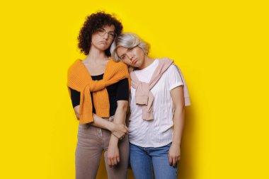 Sad caucasian sisters with curly hair are posing on a yellow studio wall gesturing upset feelings with lips
