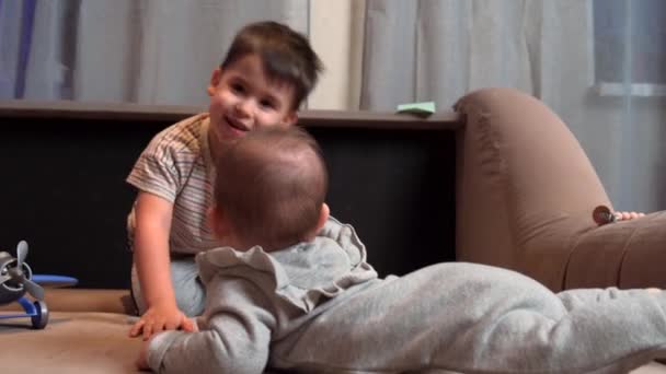 Cute caucasian boy is playing with his newborn brother kissing him while lying on the couch — Stok video