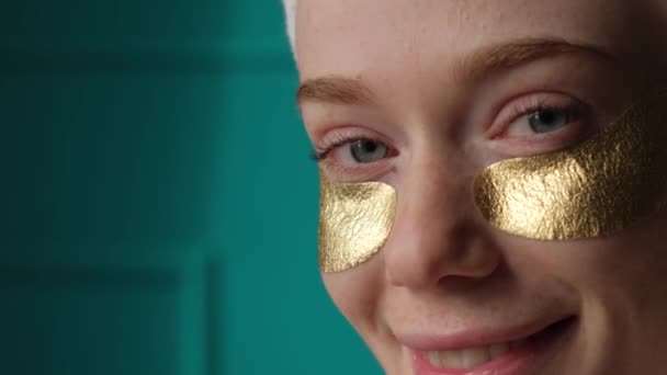 Ginger γυναίκα με χρυσά επιθέματα μάτι χαμογελώντας στην κάμερα κατά τη διάρκεια της θεραπείας του δέρματος — Αρχείο Βίντεο