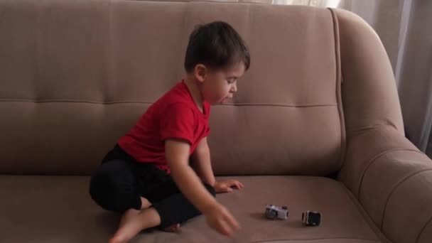 Cute caucasian boy is playing with cars on the couch simulating driving and crashing — Stockvideo