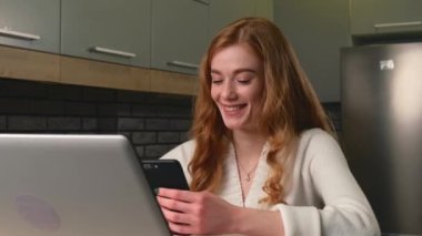 Genç girişimci mutfakta dizüstü bilgisayar kullanırken gülümsüyor ve cep telefonuyla sohbet ediyor.