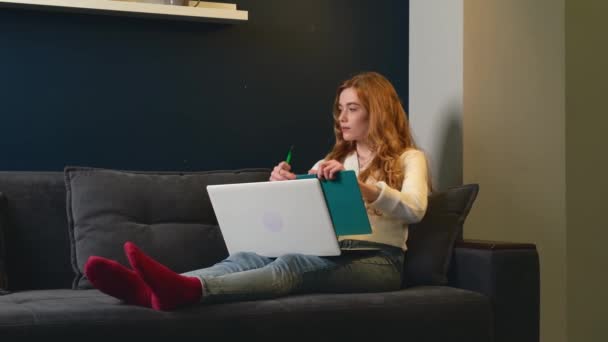 Wanita kaukasia dengan rambut merah dan bintik-bintik menulis sesuatu sambil menggunakan laptop berbaring di sofa — Stok Video