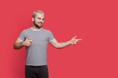Sakallı, sarışın, sevimli bir adam kırmızı bir stüdyo duvarındaki boş yeri işaret ediyor.