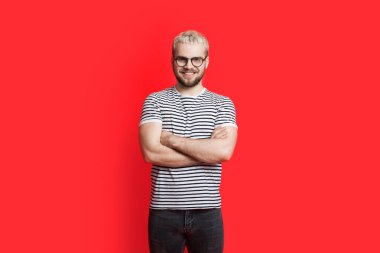 Yakışıklı adam kırmızı stüdyo duvarında ellerini çapraz yapıp kameraya gözlükleriyle gülümsüyor.