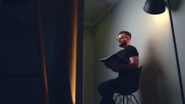 Sakallı ve gözlüklü gülümseyen adam internetten video eğitimi alıyor ve elinde stüdyo duvarında bir kitap tutuyor.