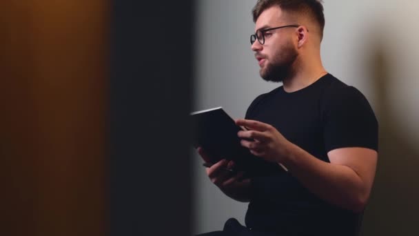 Kaukasischer Mann mit Brille und Bart liest während eines Online-Kurses von zu Hause aus ein Buch — Stockvideo