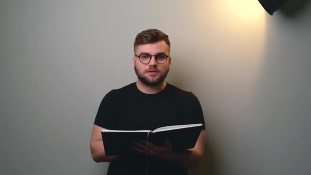 Vídeo com vista frontal de um homem caucasiano lendo um livro na frente da câmera em uma parede de estúdio cinza — Vídeo de Stock