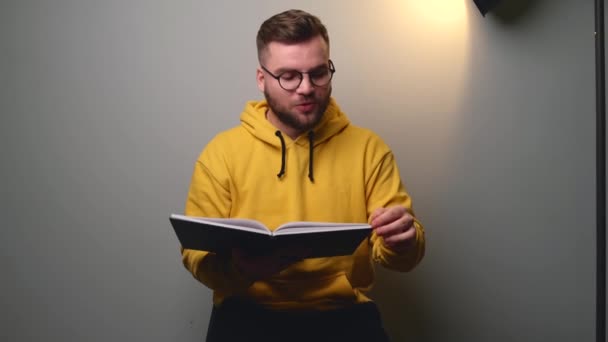 Vit man i gul tröja talar inför kameran medan han läser ur en bok på en grå studiovägg. — Stockvideo