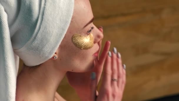 Mulher caucasiana usando uma toalha na cabeça está aplicando manchas olho dourado hidrogel — Vídeo de Stock